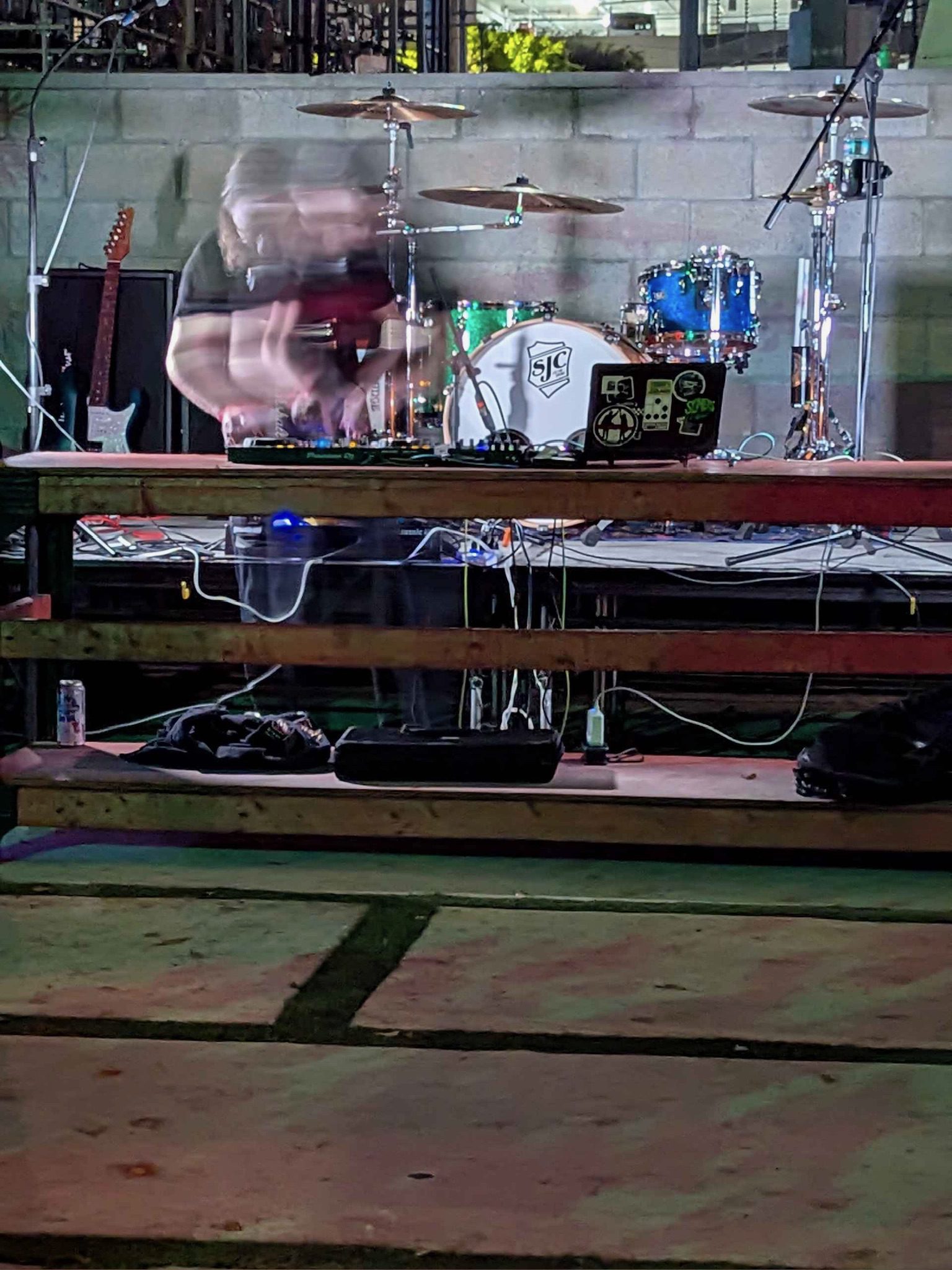 A man DJs at a big wooden table in front of a drum set, some guitars, and in the background is an industrial zone. The man is blurry; caught in motion.