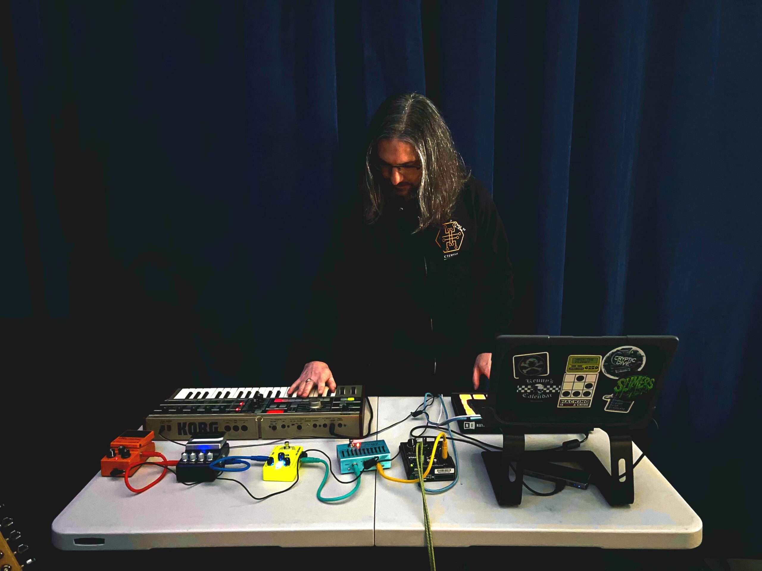 A white man with long hair in all black standing over a table of electronic music equipment.